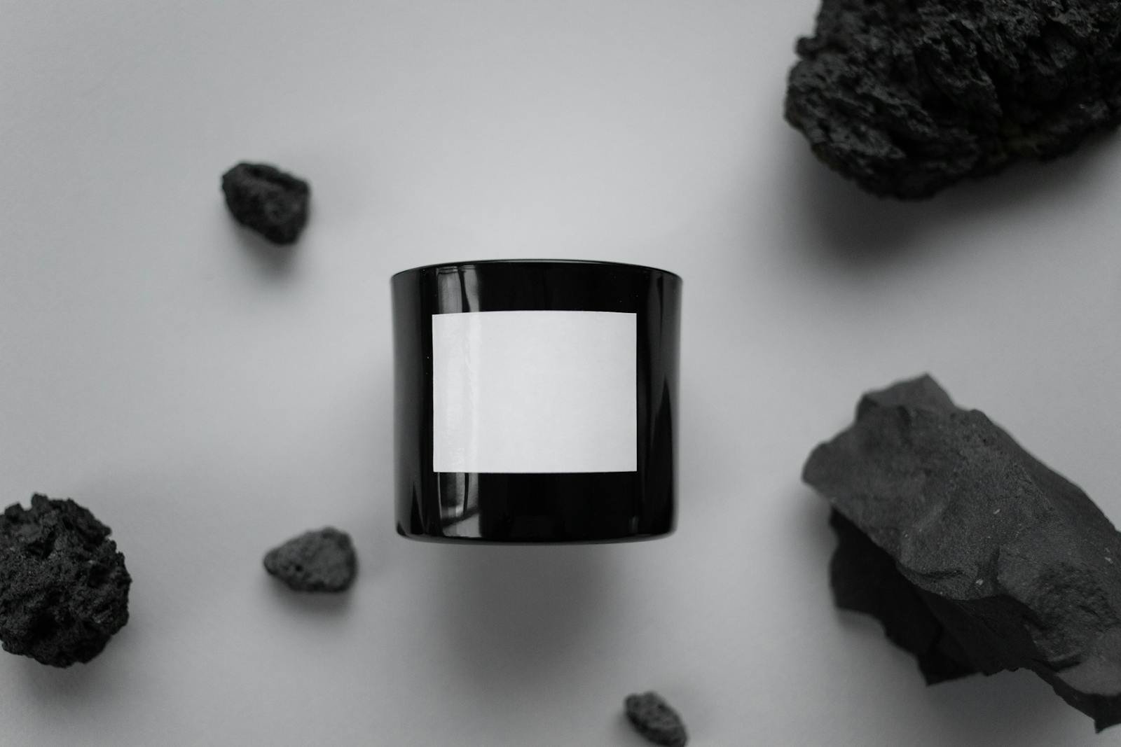 A minimalist still life featuring a black candle with a blank label surrounded by charcoal rocks.